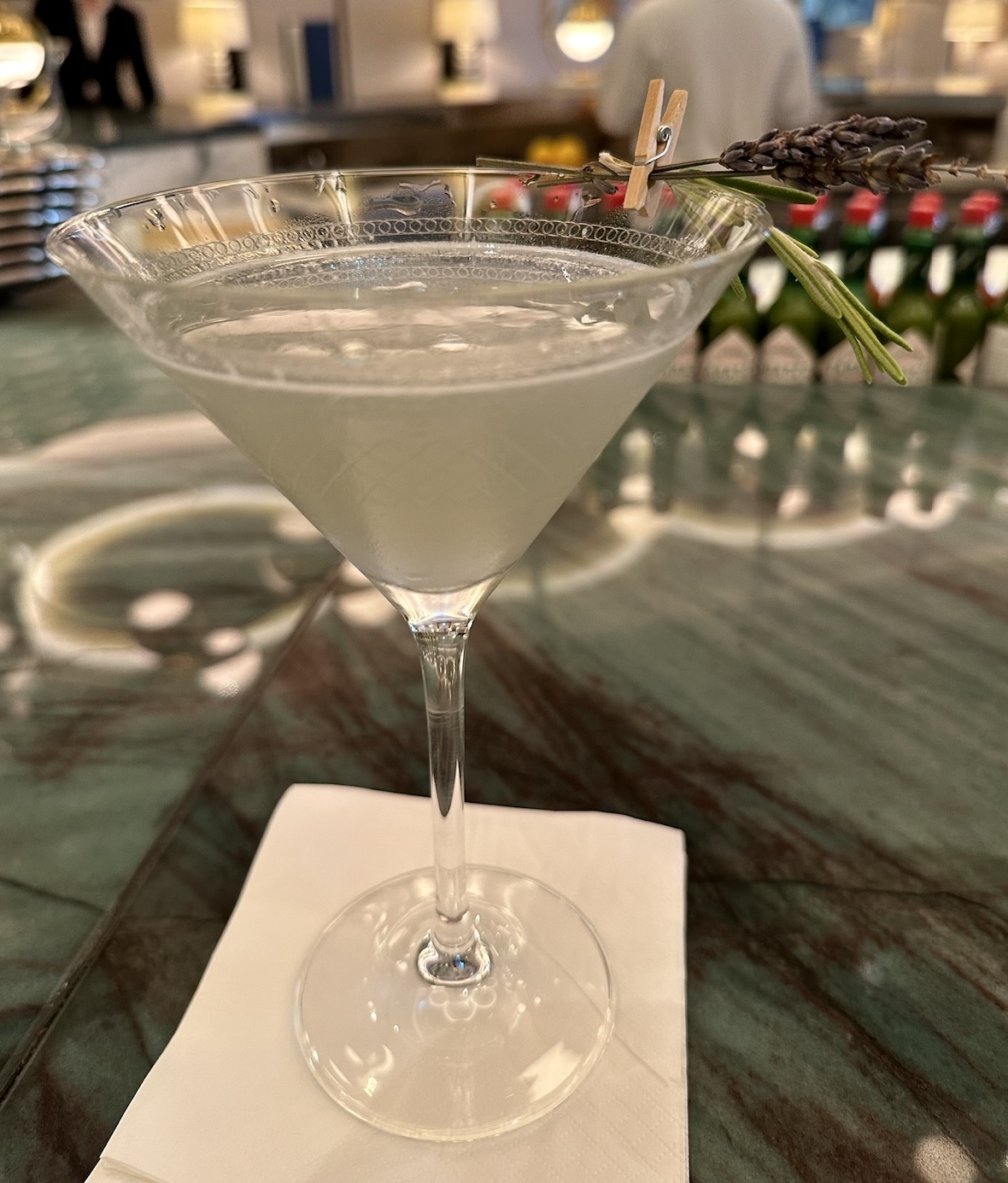 A cocktail glass on a bar counter containing a mixed drink and decorated with sprig each of lavender and rosemary.