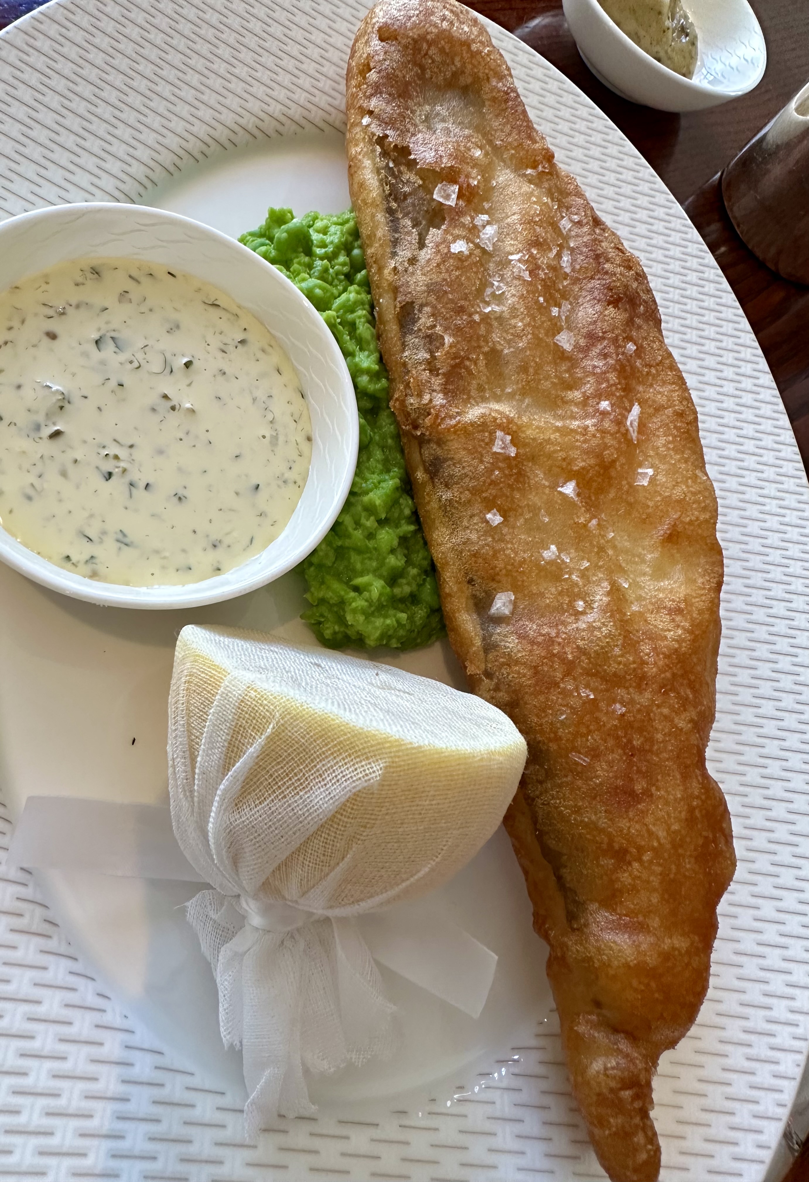 A golden brown, battered fish on a bed of crushed peas.  A small bowl of tartare sauce also sits on the plate along with a half lemon covered in tied gauze.